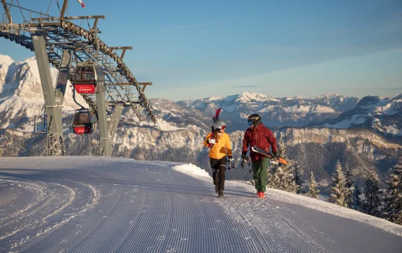 Skiers walking along next to ski lift with mountains in background